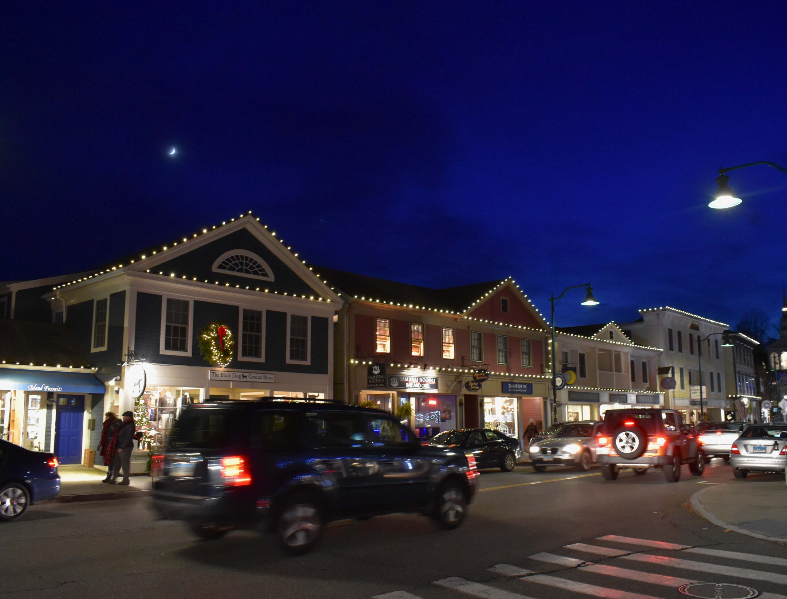 Cars drive down Main Street in Mystic on a Winter's evening while the buildings are decorated for the Holliday. 